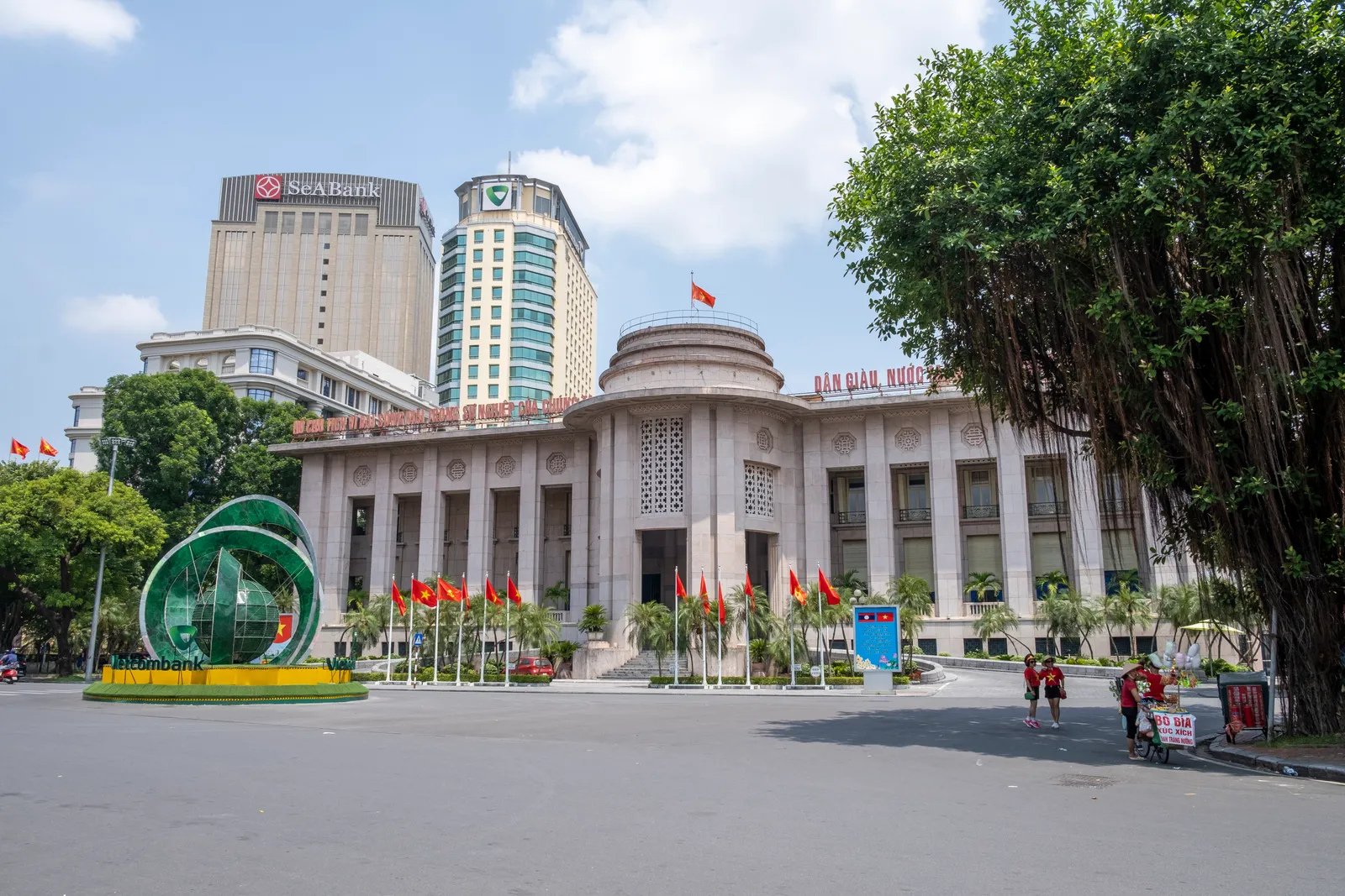 The State Bank of Vietnam headquarters in Hanoi, a pale stone building with a domed centre and a row of red flags in front, commercial bank towers behind it