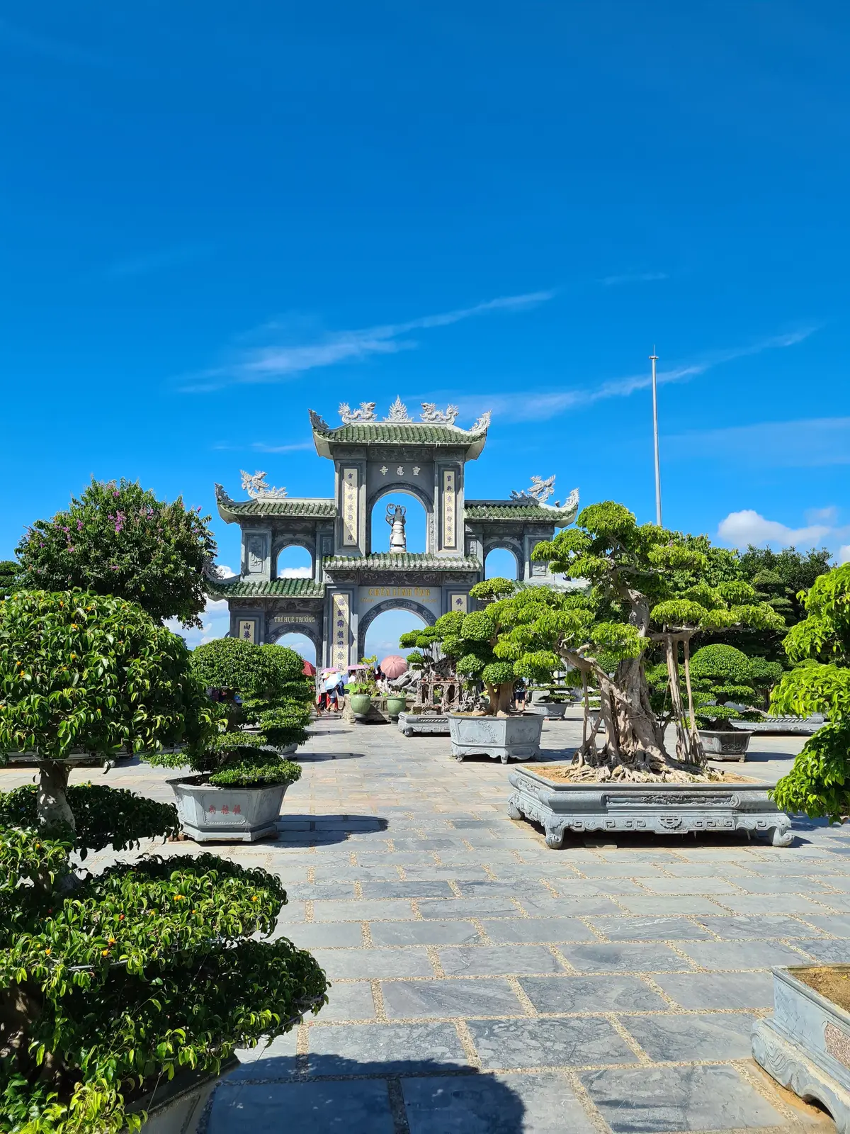 An ornate triple-arched stone gate behind rows of bonsai trees in a paved courtyard.