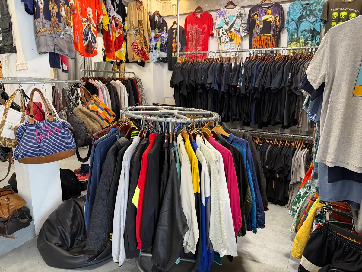 Racks of second-hand sports jackets and a wall of bags in a bright, white vintage clothing shop.