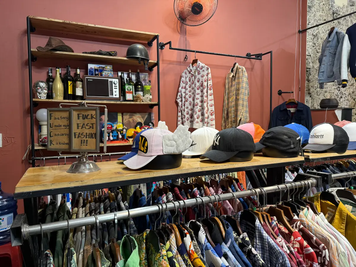 A wooden table of caps above a rail of shirts in the Nighki2hand shop, with shelves of bags and bottles behind.