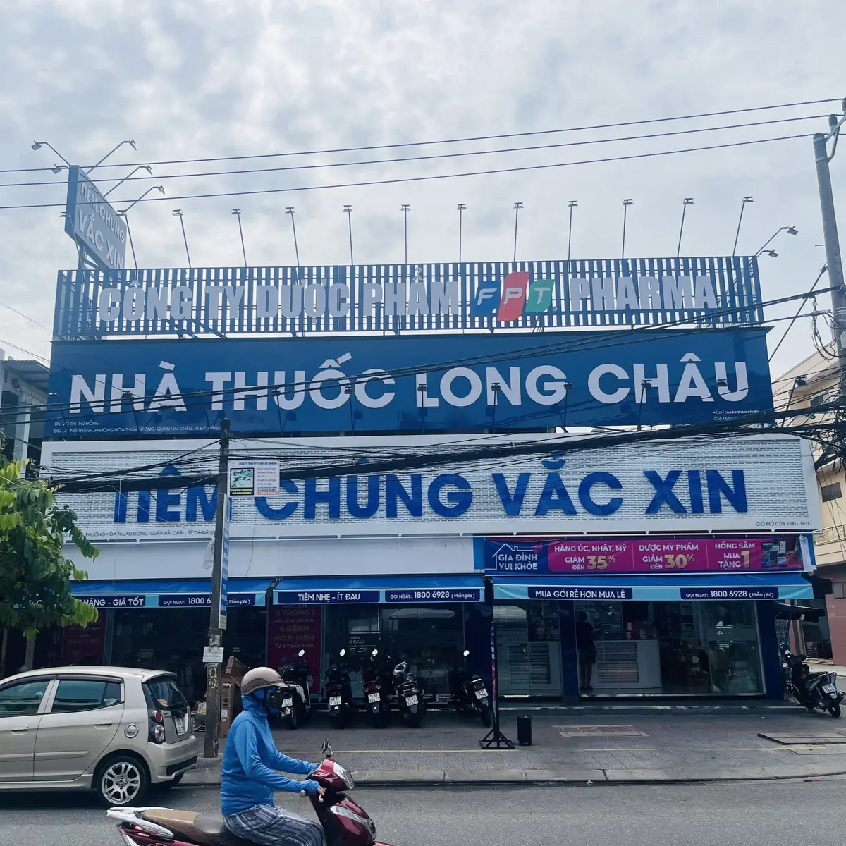 예방접종 표지판 위에 큰 파란색 Long Chau 약국 간판이 달린 길모퉁이 상가예요.