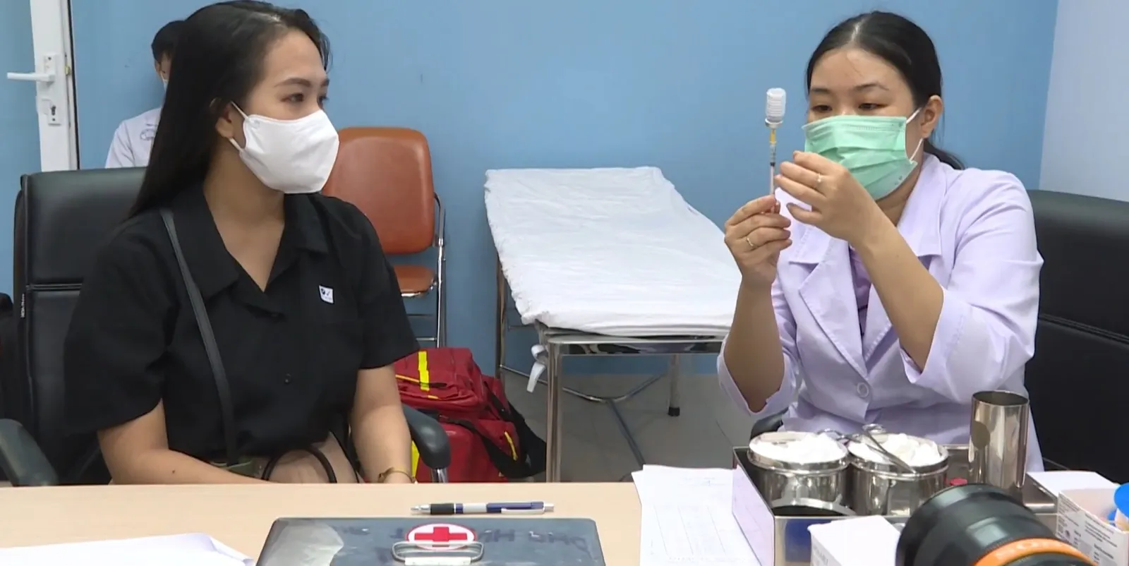 A health worker in a white coat and mask prepares a syringe at a desk while a masked woman waits beside her.