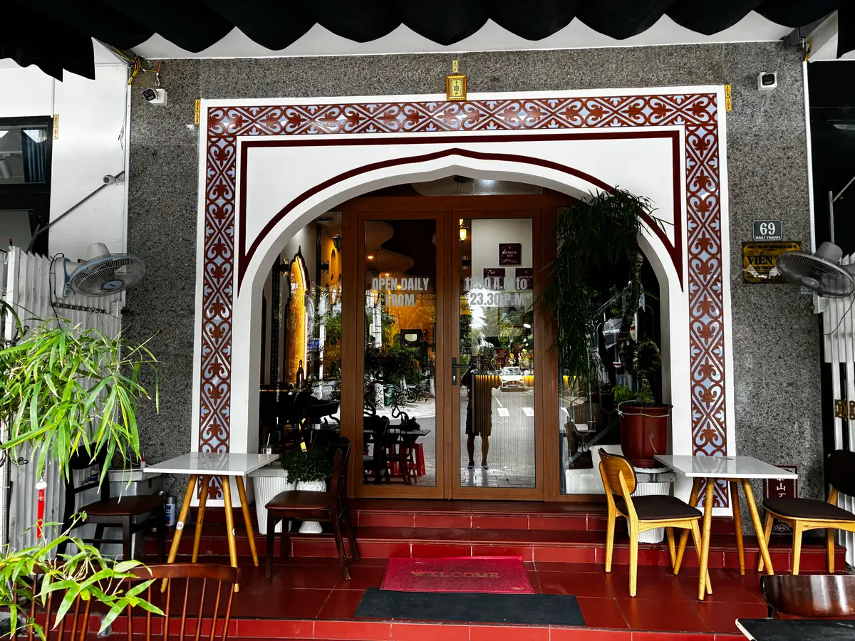 흰색과 빨간색 아치 아래 이중 나무문이 있고 왼쪽에 야외 테이블이 있는 Indian Aroma Restaurant의 입구.