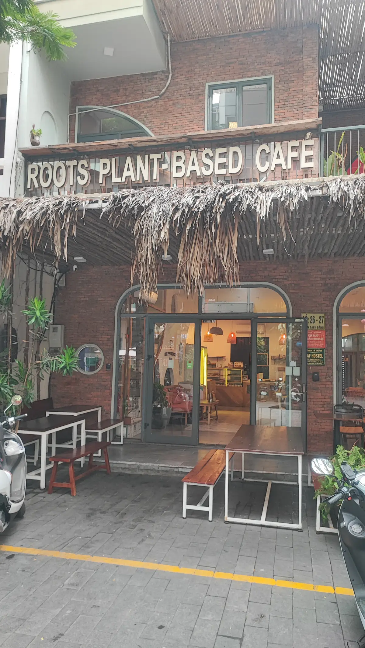 유리문, 바깥 테이블, 초가지붕 처마가 있는 ROOTS Plant-Based Cafe의 벽돌 입구.