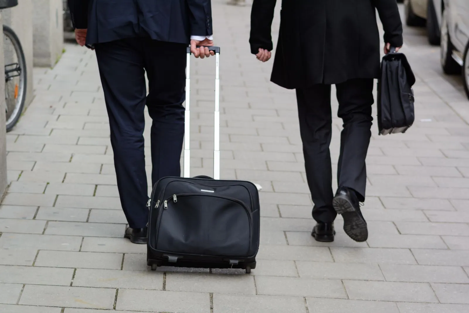 Two people in dark suits walking away down a paved street, one pulling a wheeled case and the other carrying a briefcase