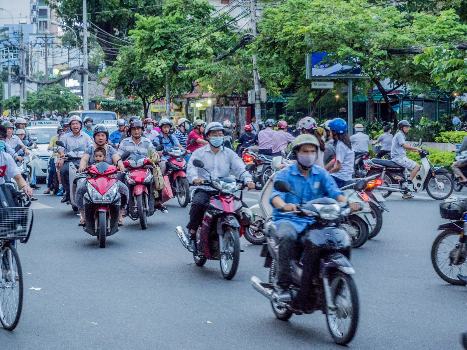 Dense motorbike traffic on a tree-lined Vietnamese city street, riders in helmets and face masks, one carrying a small child