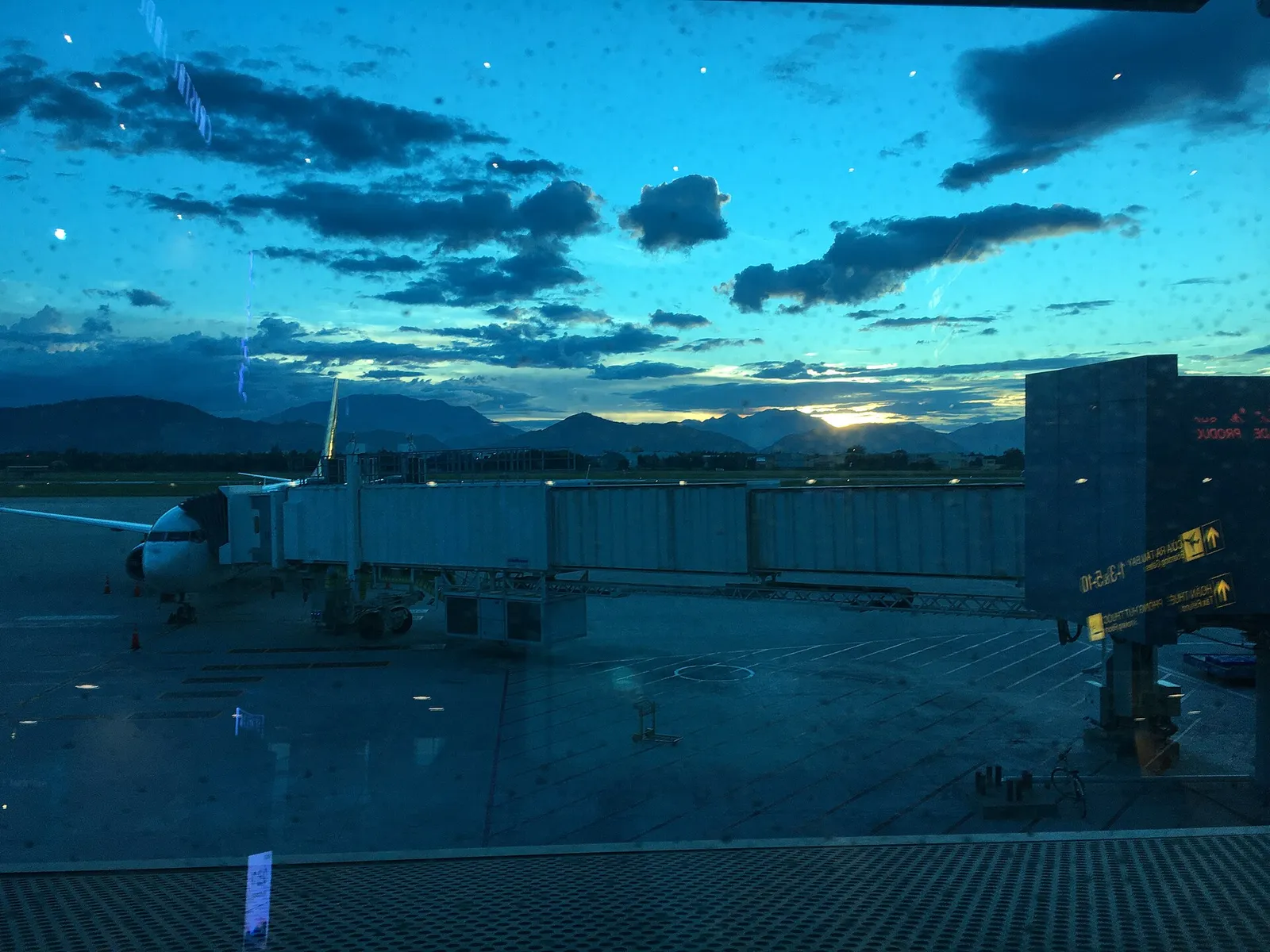 雨粒のついたターミナルのガラス越しに見る、夕暮れのダナン国際空港。駐機中の航空機につながるボーディングブリッジと、地平線上の山々が見えます。