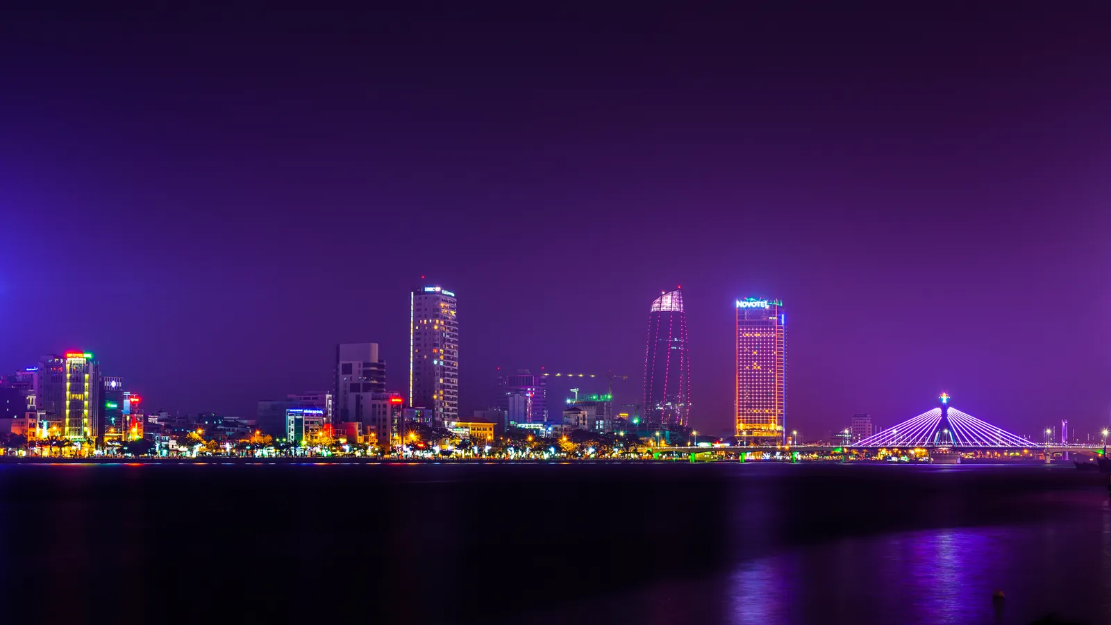 Da Nang skyline at night across the Han River, with lit high-rise towers and the illuminated cable-stayed Han River Bridge on the right