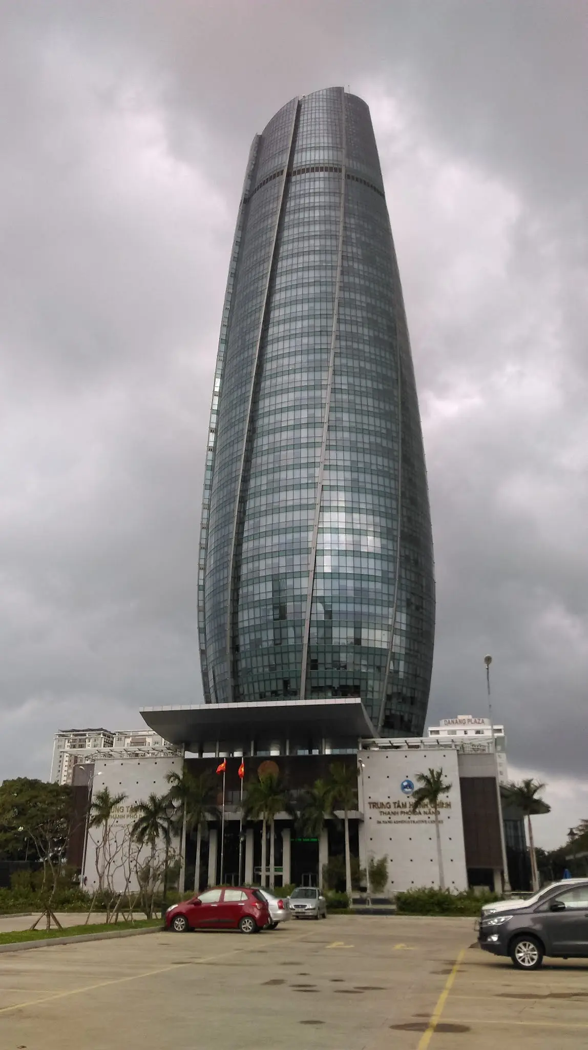一座弯曲的高大玻璃塔楼矗立在低矮的入口建筑和停车场后面，天空多云。