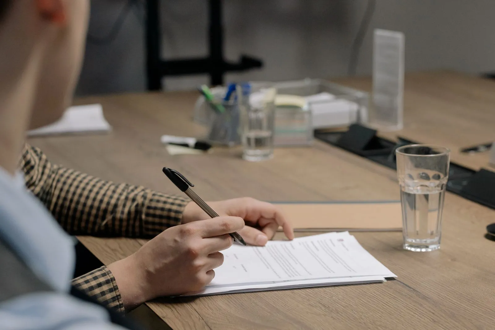 A person at an office meeting table signing a printed document, a glass of water beside it